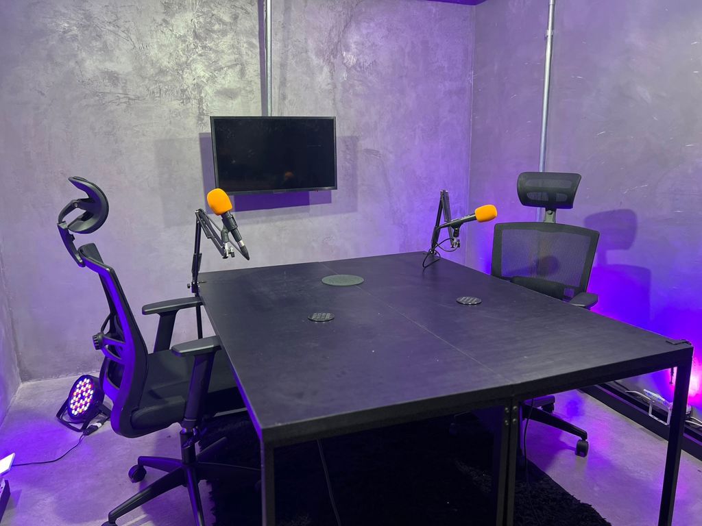 Sala que comporta 4 pessoas confortavelmente. Ambiente descontrído, cenários de podcast, com variedade de luzes disponíveis.