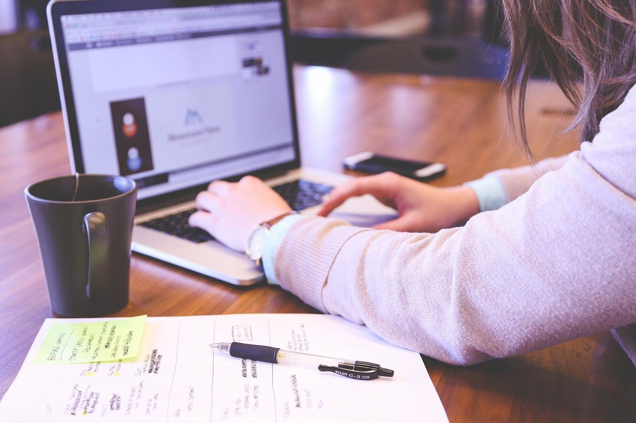 Computador sob uma mesa, com um papel com anotações e uma xícara ao lado e pessoa com as mãos no teclado.