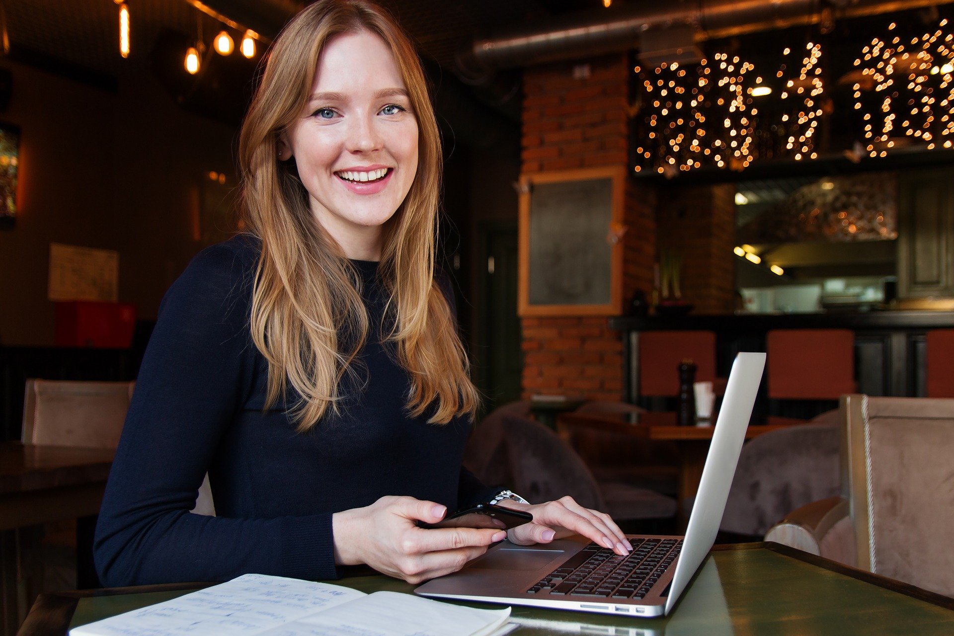 Mulher trabalha sorridente com tecnologias portáteis como celular e notebook.