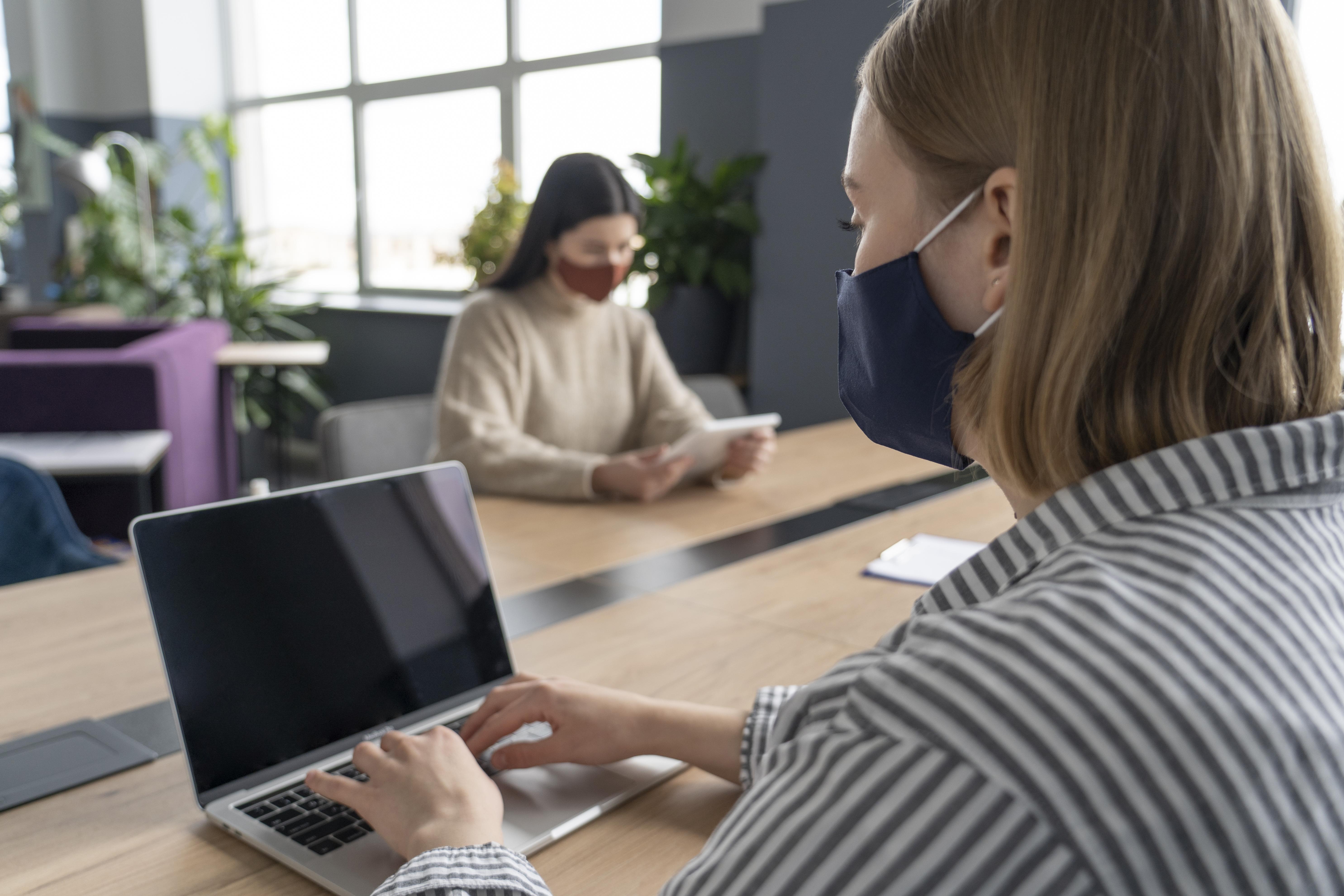 Mulher trabalha num coworking respeitando o distanciamento social e fazendo uso de máscara.